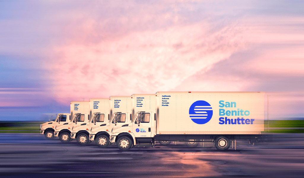 A fleet of large box trucks with San Benito Shutter logo on their sides, parked on an asphalt parking lot with futuristic styling to show the trucks are in movement.